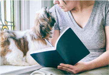 Woman taking educational course notes in a notebook while looking at her dog beside her.